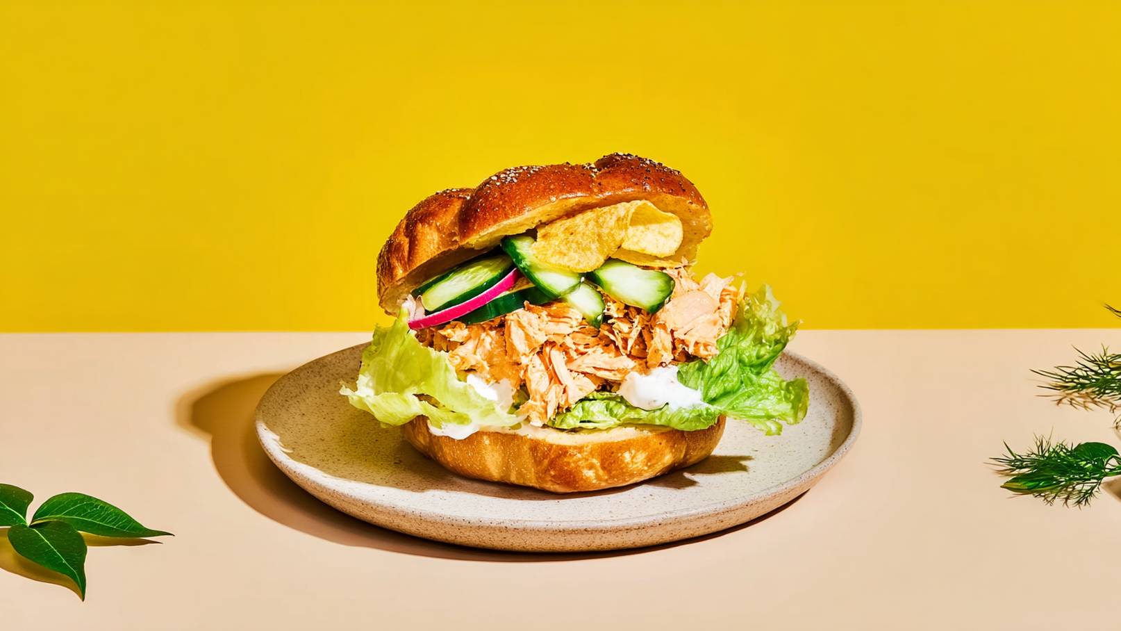 Ein saftiger Sandwich auf einem Teller, gefüllt mit Hühnchen, Salat, Gurkenscheiben und roten Zwiebeln. Das Brötchen hat eine goldbraune Farbe und ist leicht knusprig. Im Hintergrund ist eine helle, gelbe Wand sichtbar.