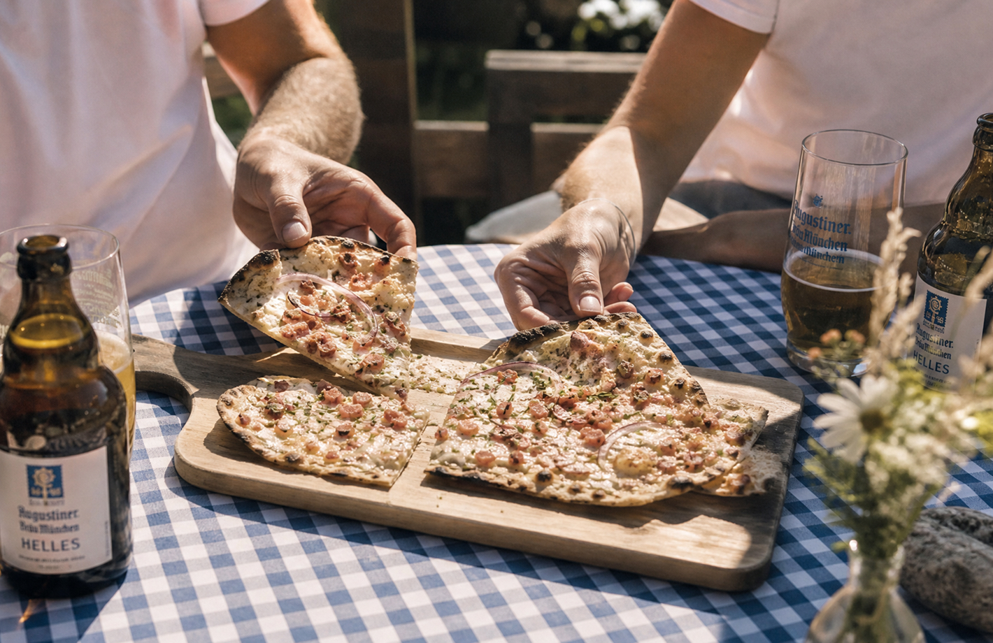 Zwei Personen teilen an einem Gartentisch eine herzförmige Pizza auf einem Holzbrett; Hände greifen Stücke, daneben Bierflaschen und -gläser.