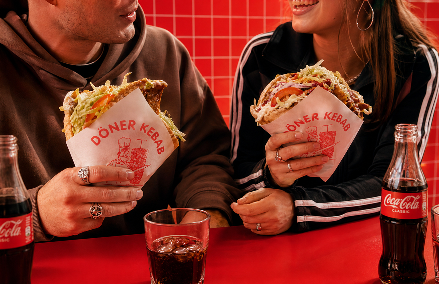 Zwei Personen sitzen an einem roten Tresen und halten jeweils einen in Papier gewickelten Döner Kebab mit Salat und Sauce; auf dem Tresen stehen eine Glasflasche Coca‑Cola und ein Glas mit Cola.
