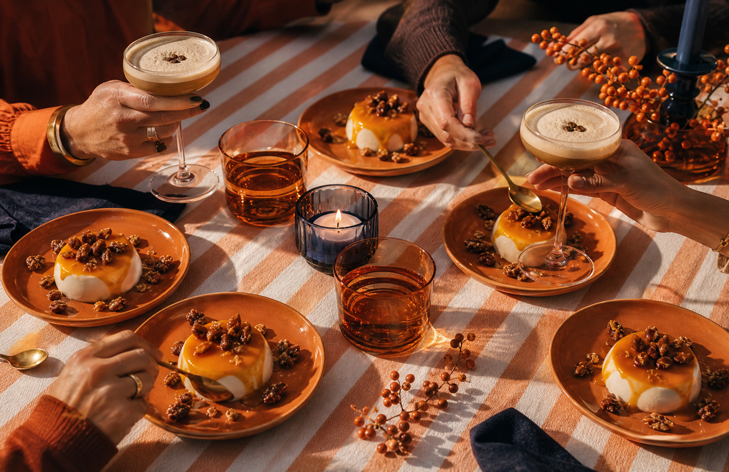 Vier Teller mit Panna‑Cotta‑ähnlichem Dessert, Karamellsauce und Nüssen auf gestreiftem Tisch; mehrere Hände mit Löffeln und Getränken, Kerze und herbstliche Dekoration in warmen Tönen.