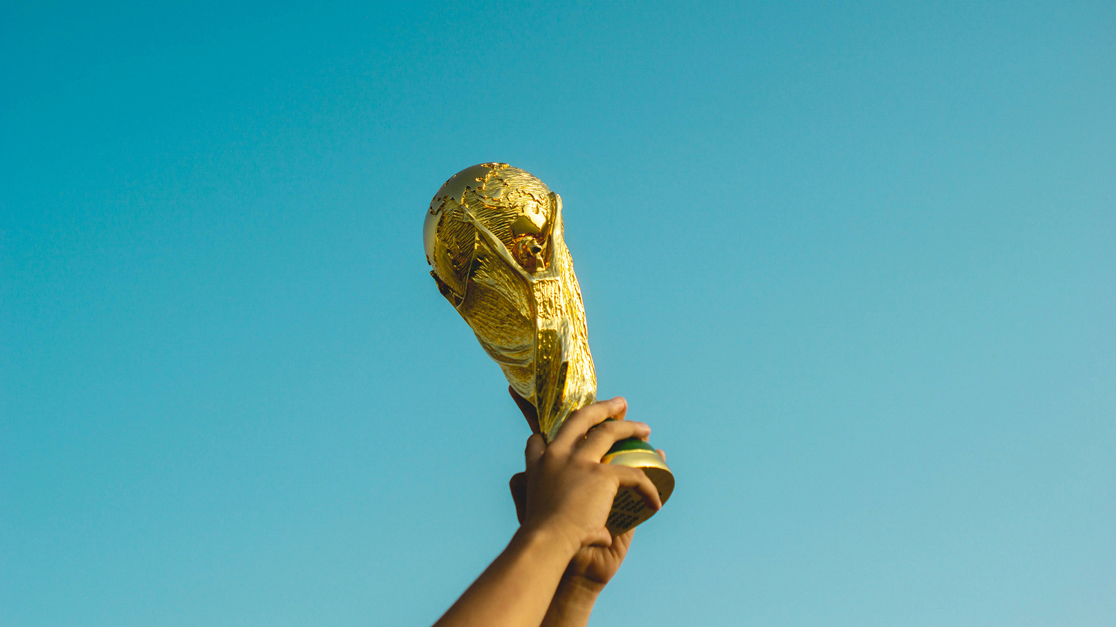 Zwei Hände halten die goldene Fußball-Weltmeisterschaftstrophäe hoch vor klarem blauem Himmel.