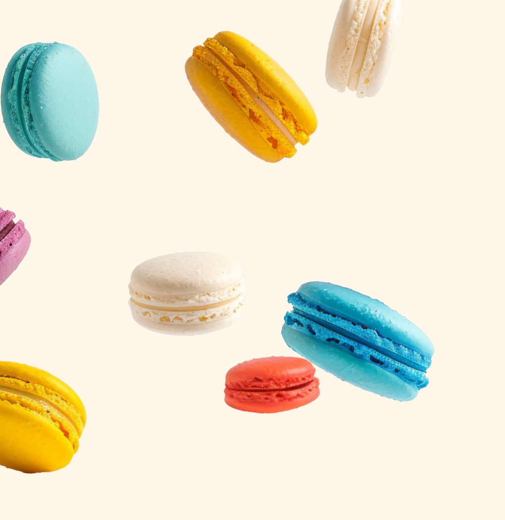 Eine Gruppe von bunten Macarons schwebt in der Luft. Die Farben umfassen Blau, Gelb, Rot, Lila und Weiß. Der Hintergrund ist hellbeige, sodass die Farben der Macarons deutlich hervortreten.
