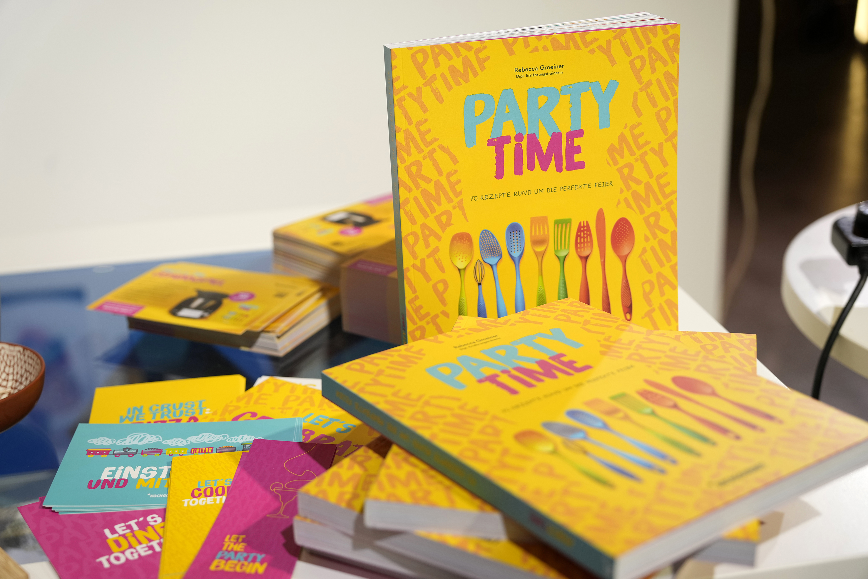 Auf dem Bild sind mehrere Kochbücher mit dem Titel „PARTY TIME“ zu sehen. Das Buch bietet 70 Rezepte für eine perfekte Feier. Der Hintergrund ist gelb und enthält bunte Utensilien wie Löffel und Gabeln. Weitere bunte Materialien sind im Vordergrund angeordnet.