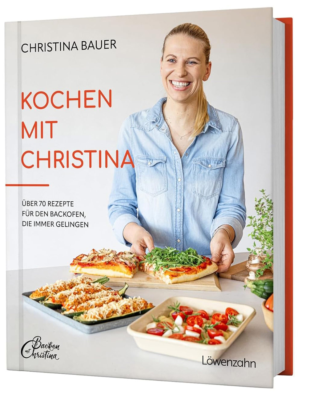 Auf dem Cover des Kochbuchs "Kochen mit Christina" lächelt eine Frau in einer blauen Jeansjacke. Sie hält zwei Pizzas mit frischen Zutaten. Im Hintergrund sind verschiedene Gerichte auf einem Tisch zu sehen. Das Buch enthält über 70 Backrezepte.