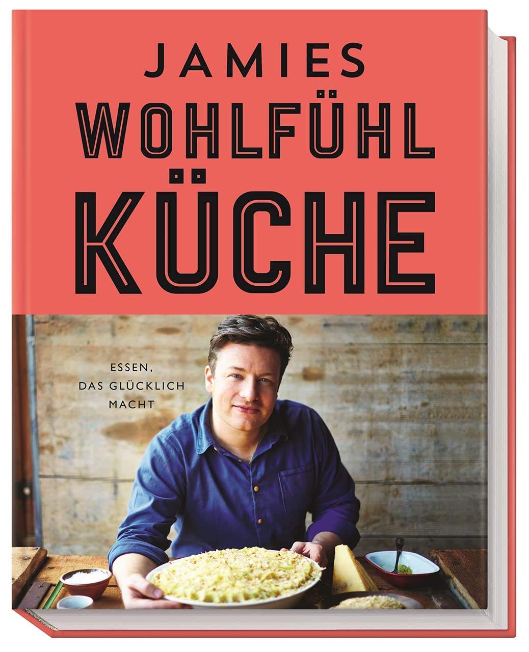 Ein Kochbuch mit dem Titel "Jamies Wohlfühlküche" auf einem rosanen Hintergrund. Ein Mann sitzt an einem Tisch und hält eine große Schüssel mit einem Gericht, umgeben von verschiedenen Zutaten. Unter dem Titel steht: "Essen, das glücklich macht."