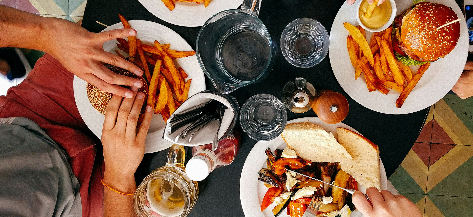 Auf einem Tisch liegen mehrere Teller mit Essen: ein Burger mit Sesambrötchen, Pommes frites, gemischtes Gemüse mit Brot und Saucen. Gläser mit Wasser und ein Krug sind ebenso sichtbar. Hände greifen nach dem Essen.