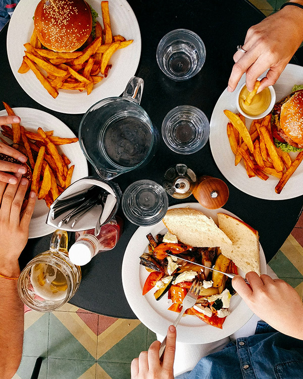 Auf einem Tisch liegen Teller mit Pommes und Burger. Ein Teller mit gegrilltem Gemüse und Brot wird mit einer Gabel und einem Messer serviert. Mehrere Gläser Wasser und ein Krug stehen bereit. Hände greifen nach dem Essen.