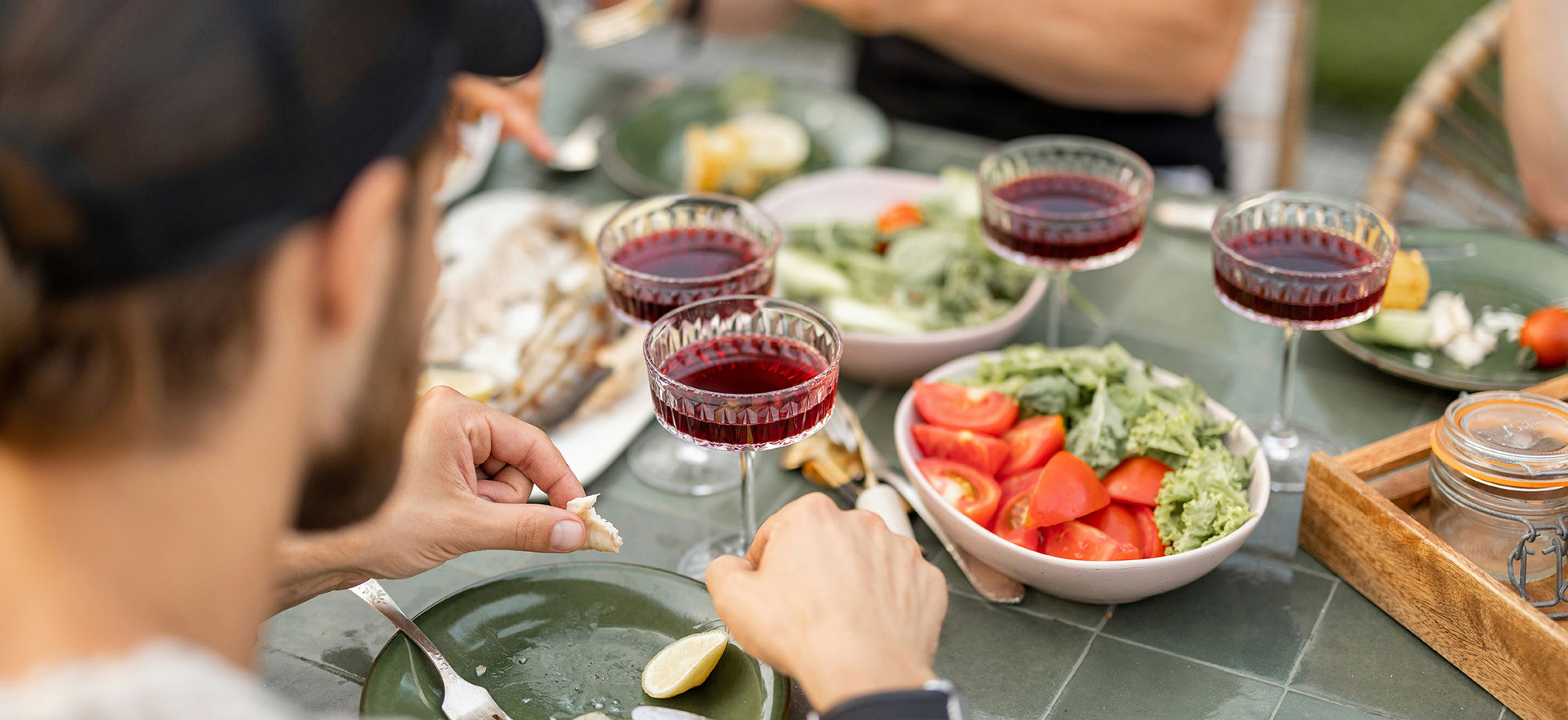 Auf dem Tisch liegt eine festliche Mahlzeit mit mehreren Salatschalen, frischen Tomaten und Gläsern mit rotem Getränk. Eine Person hält ein Stück Fisch in der Hand und trägt einen Hut, die Atmosphäre ist gesellig und einladend.