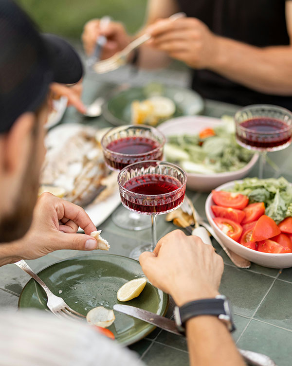 Am Tisch sind mehrere Personen versammelt, die ein Essen genießen. Es gibt Fisch, Salate und Tomaten auf den Tellern. Gläser mit rotem Getränk stehen bereit. Ein paar Personen halten Besteck und scheinen das Essen zu teilen.