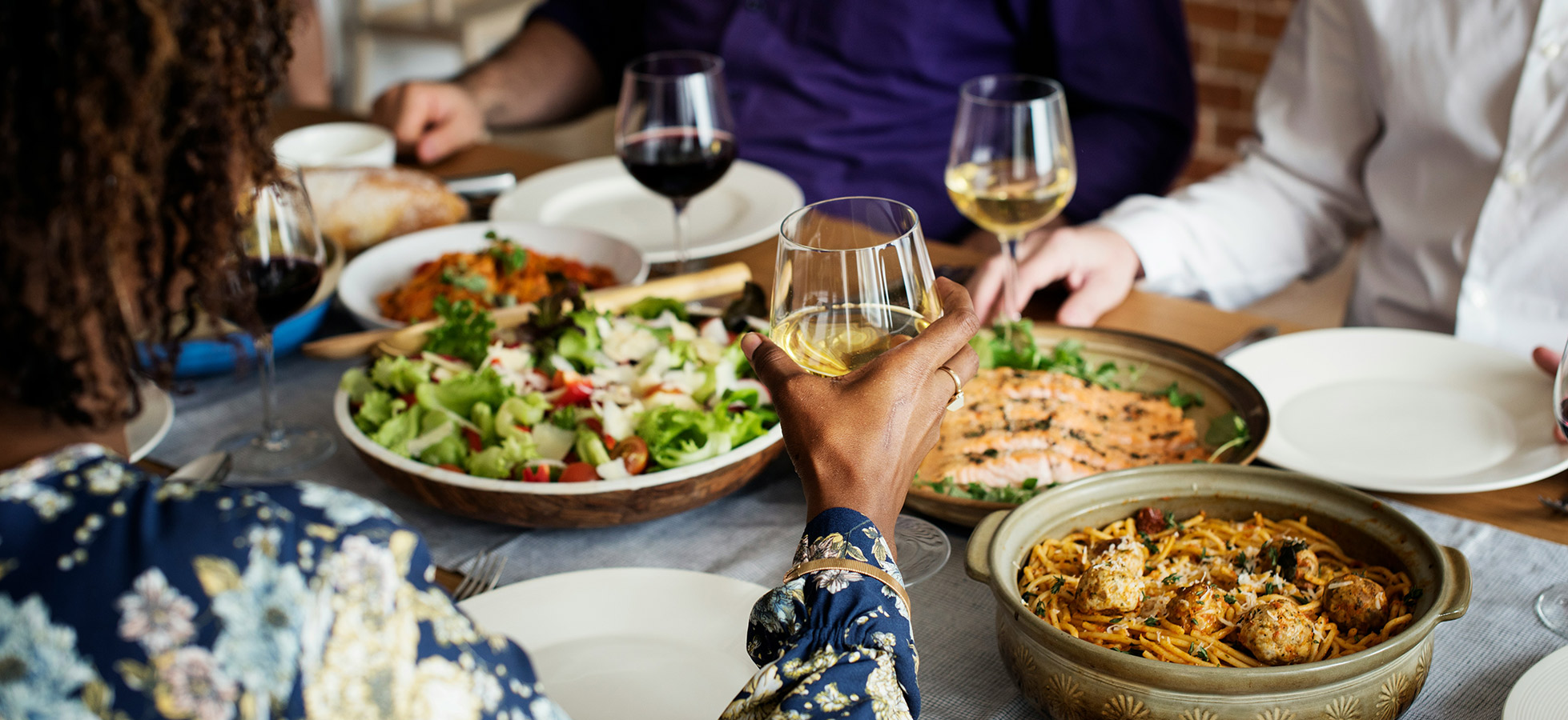 Eine Gruppe von Personen sitzt am Tisch und genießt eine Mahlzeit. Eine Hand hält ein Glas mit Weißwein, während auf dem Tisch eine Auswahl an Gerichten wie Salat, Pasta und Fisch serviert wird. Mehrere Weingläser sind ebenfalls sichtbar.