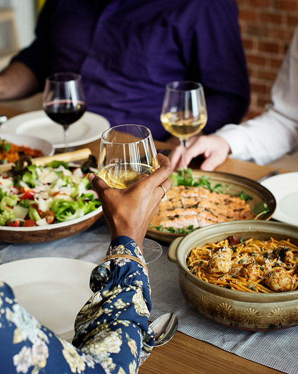 Ein gedeckter Tisch mit verschiedenen Gerichten, darunter Salat, Pasta und Lachs. Eine Hand hält ein Glas Weißwein hoch. Mehrere Personen sind am Tisch versammelt, ihre Gesichter sind teilweise sichtbar. Die Atmosphäre wirkt gesellig und einladend.