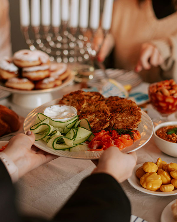 Ein Mensch hält einen Teller mit zwei knusprigen Kartoffelpuffern, frischen Gurkenscheiben, Tomaten und einem kleinen Becher Sauerrahm. Im Hintergrund sind weitere Speisen und ein Menora aus Kerzen sichtbar.