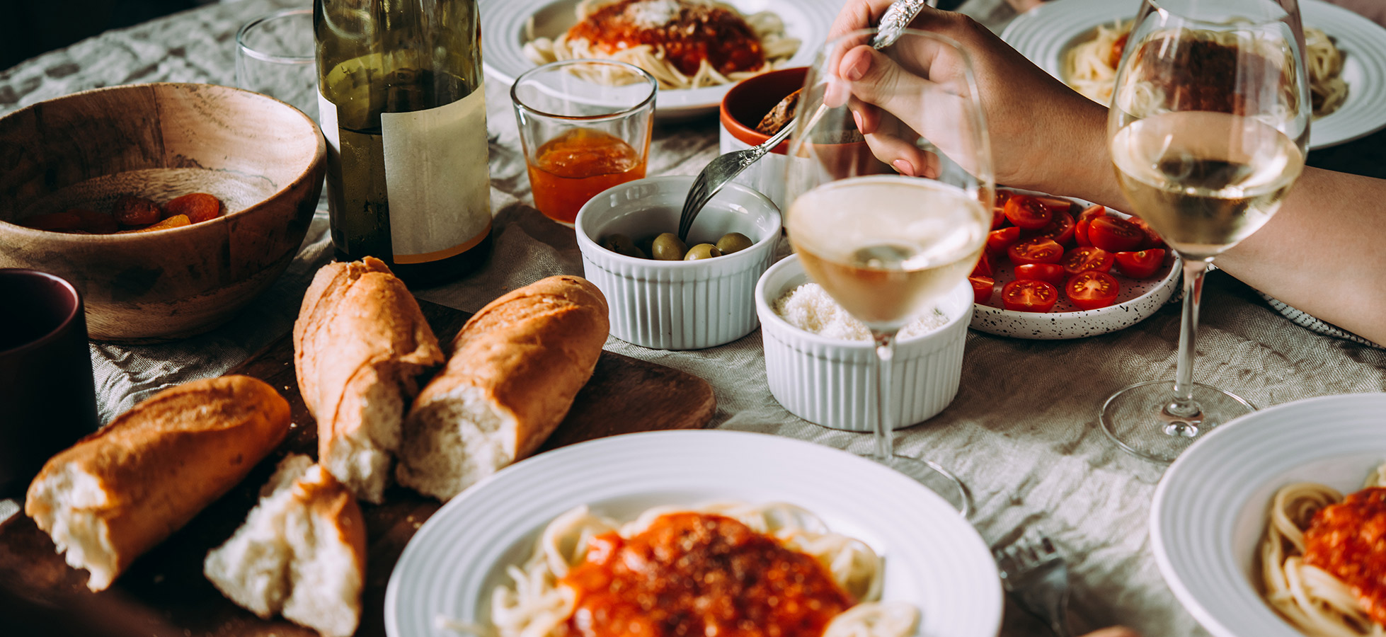 Ein Tisch ist festlich gedeckt mit verschiedenen Speisen, darunter Spaghetti mit Tomatensauce, frisches Brot, Oliven und Kirschtomaten. Es sind Gläser Wein und Saft vorhanden, sowie eine Holzschüssel mit Trockenfrüchten, die die gesellige Atmosphäre betonen.