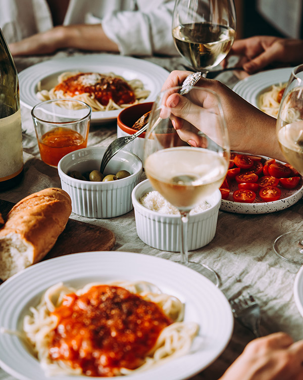 Eine gedeckte Tafel mit verschiedenen Speisen: Spaghetti mit Tomatensoße, eine Schüssel Oliven, frische Tomatenscheiben und ein Stück Brot. Gläser mit Weißwein und anderen Getränken sind ebenfalls auf dem Tisch. Hände greifen nach dem Essen, das eine gesellige Atmosphäre vermittelt.