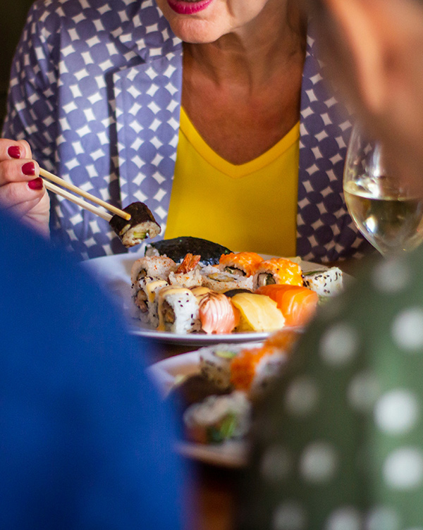 Eine Person mit einer gelben Bluse und einem gemusterten Blazer hält mit Stäbchen Sushi. Auf dem Tisch liegt ein Teller mit verschiedenen Sushi-Sorten. Im Hintergrund sind unscharf weitere Personen zu sehen und ein Glas Weißwein.