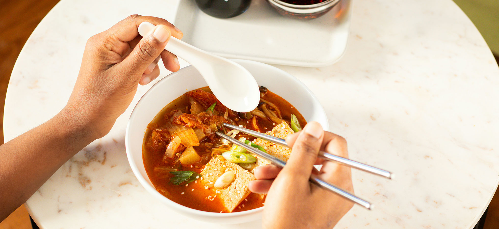 Eine Person hat einen Löffel in der einen Hand und Essstäbchen in der anderen Hand. Sie essen aus einer Schüssel mit einer würzigen Suppe, die Gemüse und Tofu enthält. Die Schüssel steht auf einem Tisch mit einer hellen Oberfläche.