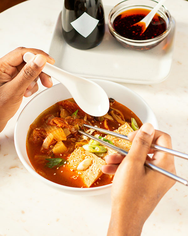 Ein Paar Hände hält Essstäbchen und einen Löffel über eine Schüssel mit würziger Suppe. Die Suppe enthält Tofu, Gemüse und Gewürze. Im Hintergrund sind Sojasauce und eine Schüssel mit rotem Dip zu sehen.