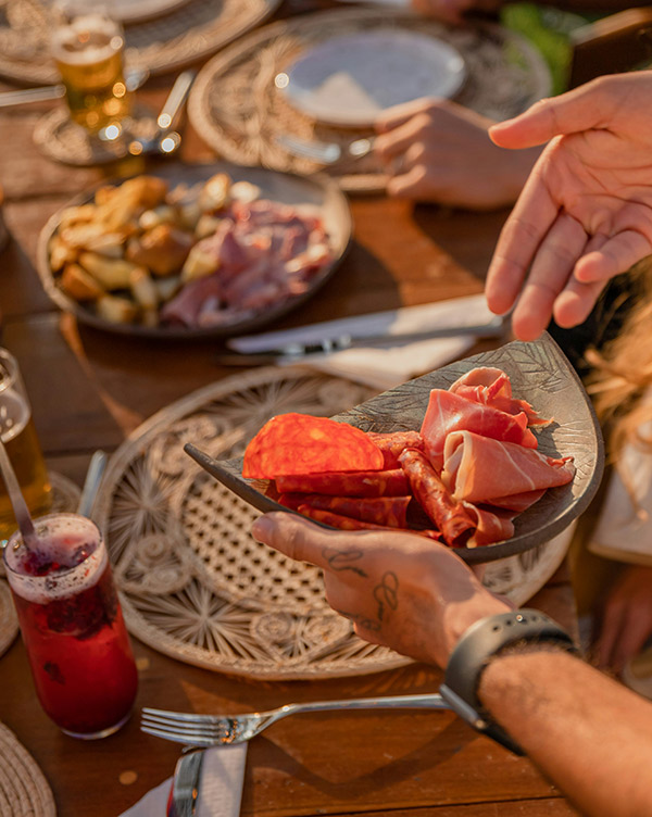 Ein Teller mit verschiedenen Wurstsorten wird von einer Person über den Tisch gehalten. Im Hintergrund sind weitere Teller mit Essen und Getränke zu sehen. Die Szene vermittelt eine gesellige Atmosphäre beim gemeinsamen Essen.