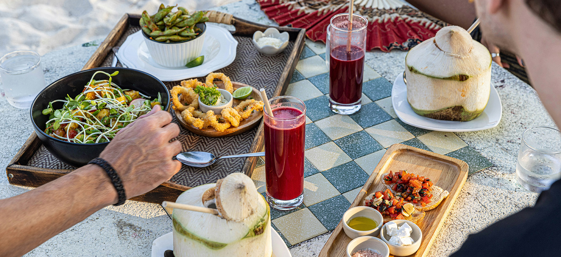 Auf einem Tisch in einem Freiluftbereich sind verschiedene Speisen und Getränke angerichtet. Dazu gehören ein bunter Salat, frittierte Calamari, edamame und frische Säfte in Gläsern. Eine halbe Kokosnuss und kleine Schalen mit Dips ergänzen das Essen.