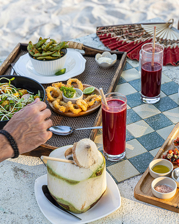Ein Tablett voller Speisen und Getränke am Strand: eine Schüssel mit Salat, knusprige Tintenfischringe, eine Schale mit Edamame und frische Limetten. Dazu zwei Gläser mit rotem Getränk und eine frische Kokosnuss mit einem Trinkhalm. Ein entspannendes Ambiente.