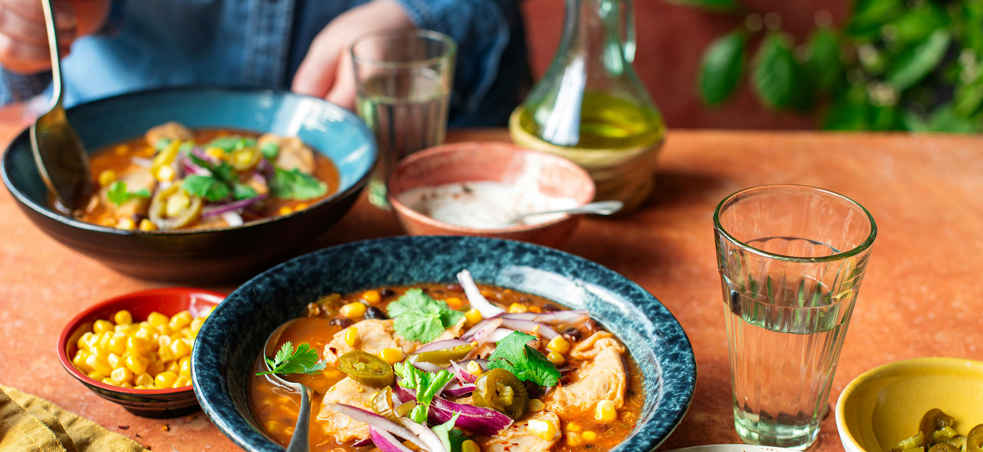 Ein Tisch mit zwei Schalen einer würzigen Suppe, die mit Hühnchen, Mais, Zwiebeln und frischem Koriander garniert ist. Daneben befinden sich kleine Schalen mit weiteren Zutaten, darunter Jalapeños und eine Portion saure Sahne. Ein Glas Wasser steht ebenfalls auf dem Tisch.