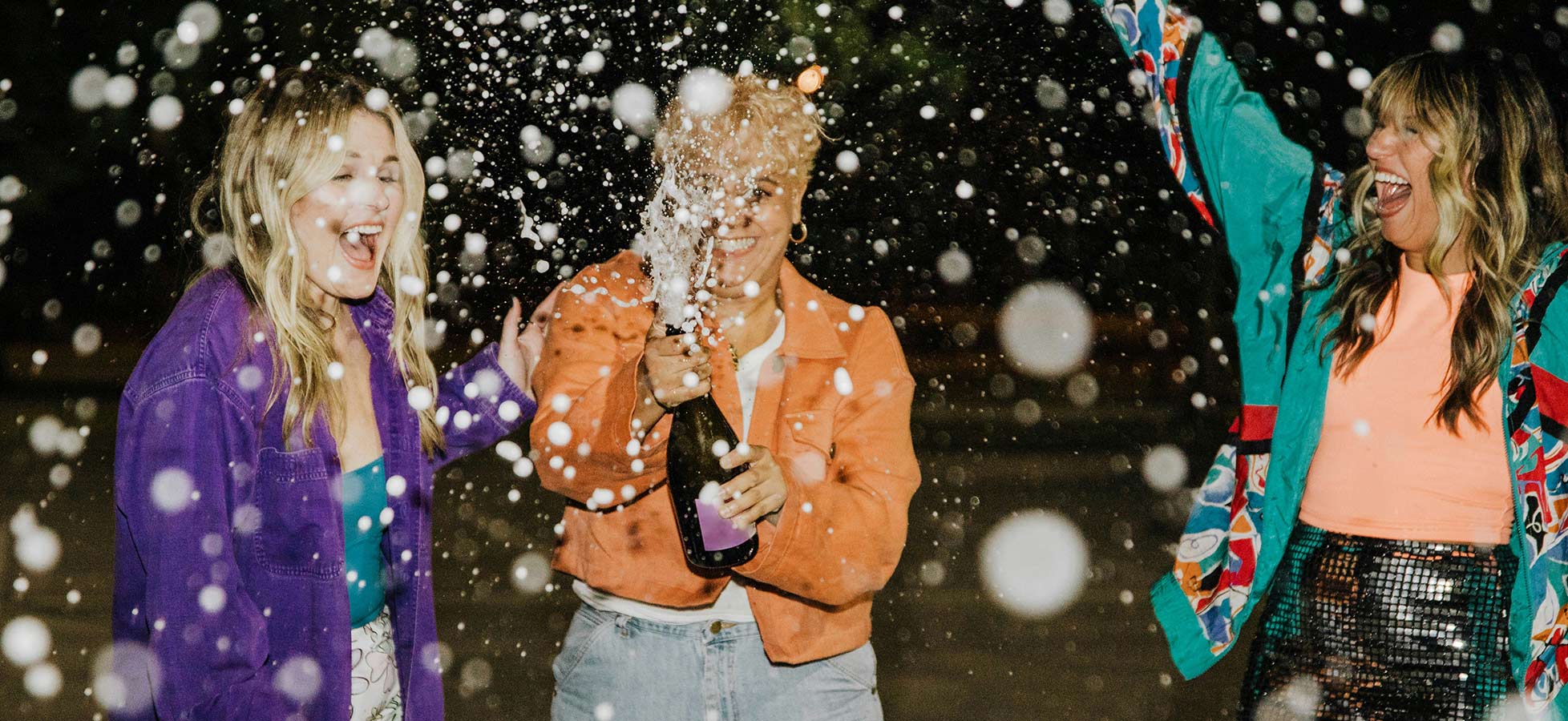 Drei Frauen feiern zusammen in der Nacht. Eine von ihnen öffnet eine Flasche Champagner, während Konfetti oder Glitzer in der Luft schwebt. Alle drei lächeln und zeigen Freude. Die Atmosphäre ist festlich und ausgelassen.
