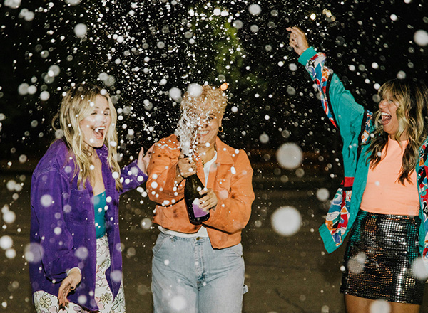 Drei Frauen feiern zusammen in der Nacht. Eine von ihnen öffnet eine Flasche Champagner, während Konfetti oder Glitzer in der Luft schwebt. Alle drei lächeln und zeigen Freude. Die Atmosphäre ist festlich und ausgelassen.