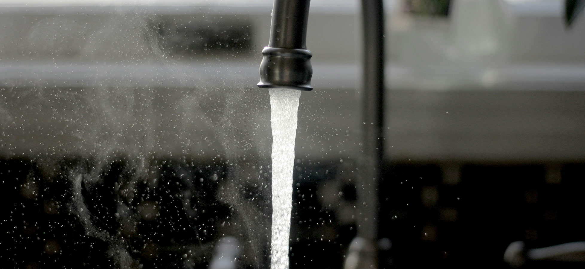 Ein Wasserhahn mit fließendem Wasser, das in einem feinen Strahl aus dem Auslauf kommt. Der Wasserstrahl erzeugt kleine Spritzer und Dampf ist sichtbar. Der Hintergrund ist unscharf, was den Fokus auf das Wasser lenkt.