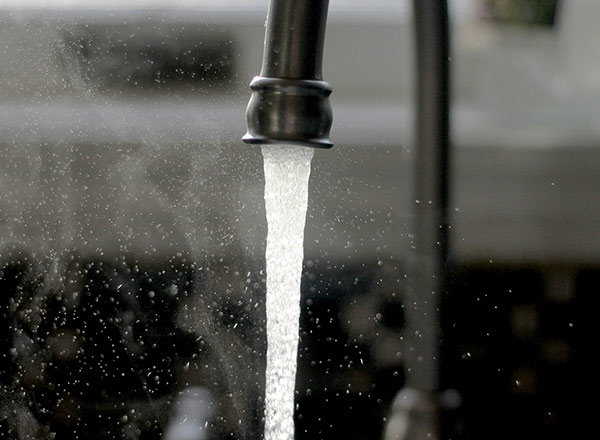 Ein Wasserhahn mit fließendem Wasser, das in einem feinen Strahl aus dem Auslauf kommt. Der Wasserstrahl erzeugt kleine Spritzer und Dampf ist sichtbar. Der Hintergrund ist unscharf, was den Fokus auf das Wasser lenkt.