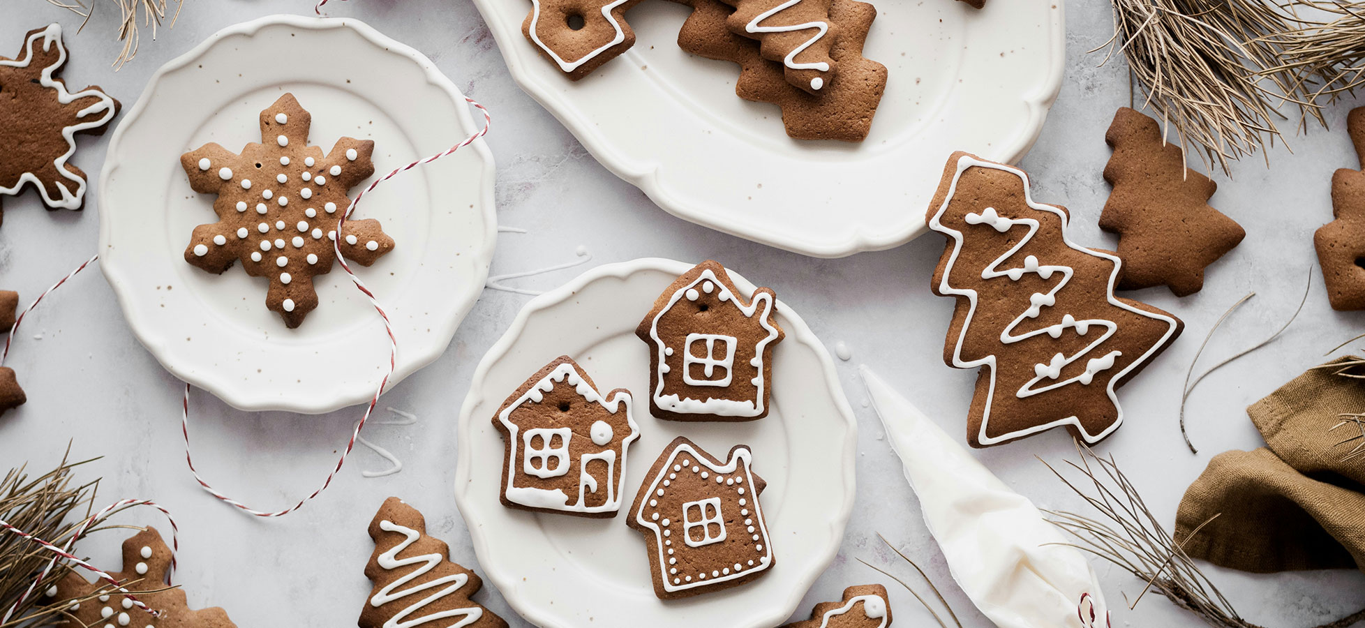Auf einem weißen Teller liegen verschiedene Weihnachtsplätzchen in Form von Schneeflocken, Tannenbäumen und Häusern, verziert mit weißem Zuckerguss. Im Hintergrund sind weitere Plätzchen und Dekorationselemente zu sehen, die eine festliche Stimmung erzeugen.