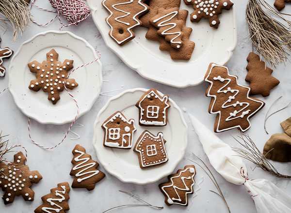 Auf einem weißen Teller liegen verschiedene Weihnachtsplätzchen in Form von Schneeflocken, Tannenbäumen und Häusern, verziert mit weißem Zuckerguss. Im Hintergrund sind weitere Plätzchen und Dekorationselemente zu sehen, die eine festliche Stimmung erzeugen.