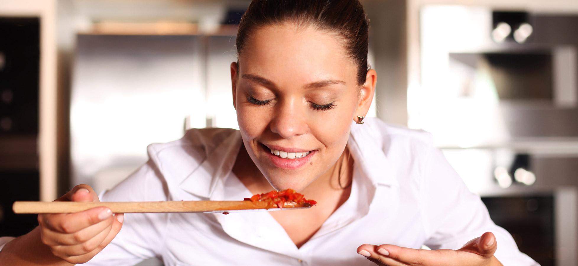 Eine lächelnde Frau in einer Küche hält einen Holzlöffel mit einem Bissen Essen. Sie schließt die Augen und genießt den Duft des Gerichts, während sie sich auf den Geschmack konzentriert.