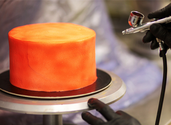 Ein runder Kuchen mit einer leuchtend orangefarbenen Oberfläche steht auf einem drehbaren Tisch. Eine Person in schwarzer Handschuhen benutzt eine Sprühpistole, um die Oberfläche des Kuchens weiter zu bearbeiten. Der Hintergrund ist unscharf und weniger wichtig für das Hauptmotiv.