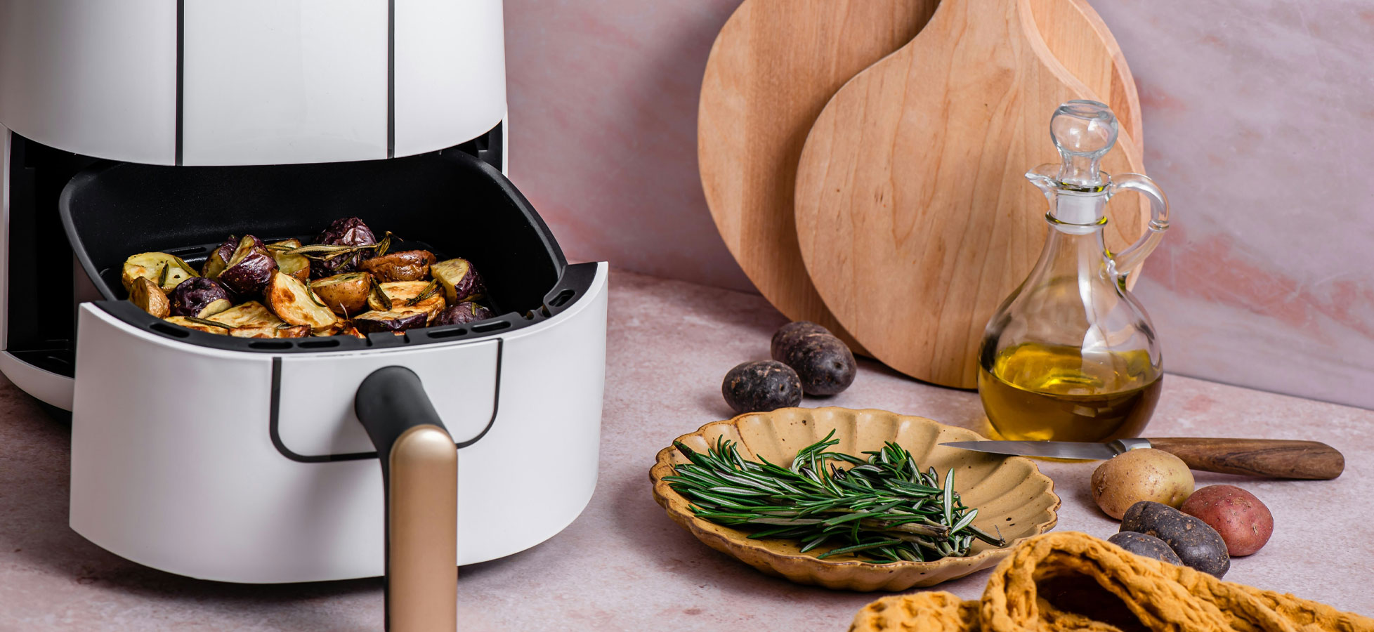 Ein moderner Heißluftfryer mit garendem Gemüse, darunter Kartoffeln und Auberginen, steht auf einer Arbeitsfläche. Rechts liegt ein Teller mit frischen Kräutern und Kartoffeln sowie eine Glasflasche mit Öl. Im Hintergrund sind Holzunterlagen erkennbar.
