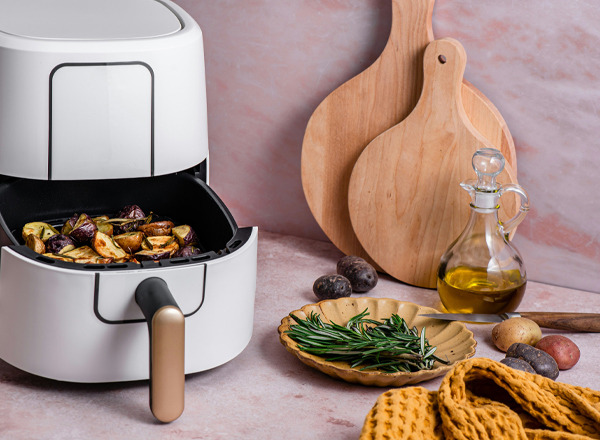 Eine Heißluftfritteuse mit geröstetem Gemüse steht auf einer Arbeitsfläche. Daneben befinden sich frische Kräuter, Kartoffeln und eine Flasche Olivenöl. Holzbrettchen und ein gelbes Küchentuch vervollständigen die Szene und vermitteln eine gemütliche Kochatmosphäre.