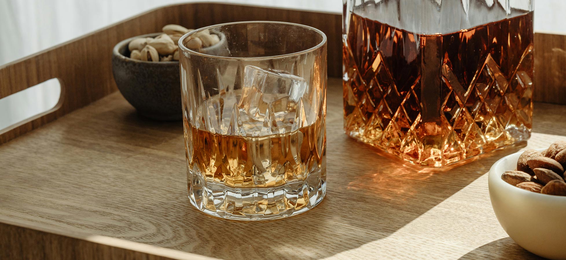 Ein Eleganter Holztablett enthält ein Glas mit einem alkoholischen Getränk, neben einer Glaskaraffe. Zudem sind kleine Schalen mit Nüssen und Mandeln sichtbar. Die Lichteffekte betonen die Farben des Getränks und das Design des Glases.