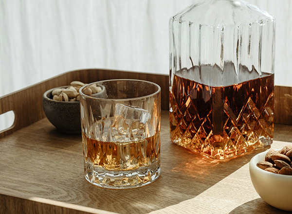 Ein Eleganter Holztablett enthält ein Glas mit einem alkoholischen Getränk, neben einer Glaskaraffe. Zudem sind kleine Schalen mit Nüssen und Mandeln sichtbar. Die Lichteffekte betonen die Farben des Getränks und das Design des Glases.