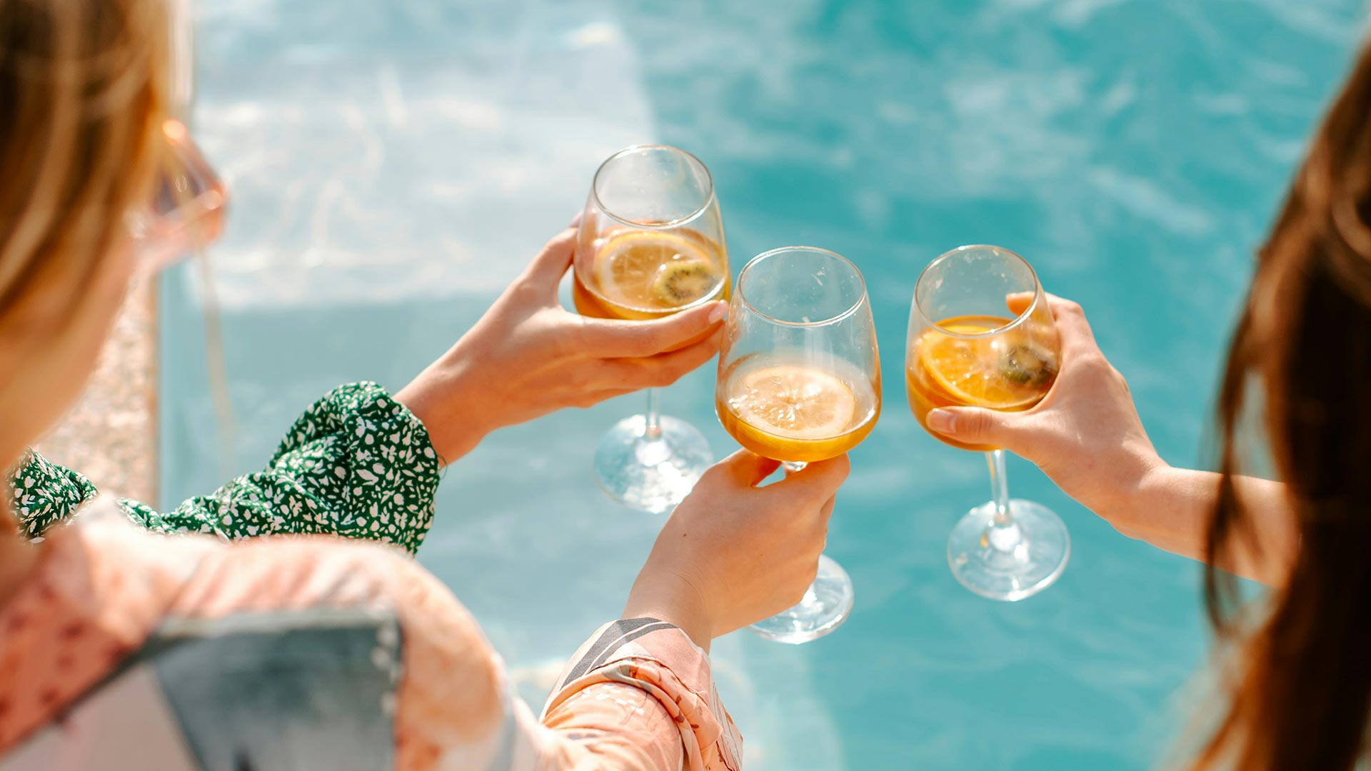 Zwei Personen stoßen mit Gläsern an, die ein Getränk mit Zitronenscheiben enthalten. Im Hintergrund ist ein blauer Pool zu sehen. Die Szene vermittelt eine fröhliche, entspannte Atmosphäre.