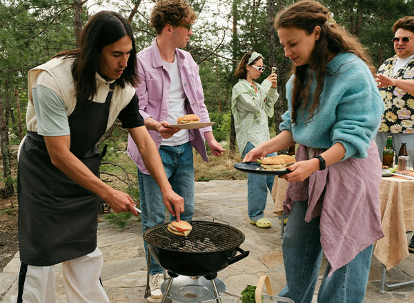 Eine Gruppe von vier Personen grillt im Freien. Ein Mann im Schürzen wendet Fleisch auf dem Grill, während eine Frau mit einem Teller zwei Burger serviert. Im Hintergrund stehen zwei Personen und eine weitere schneidet Gemüse. Alles spielt sich in einer naturnahen Umgebung ab.