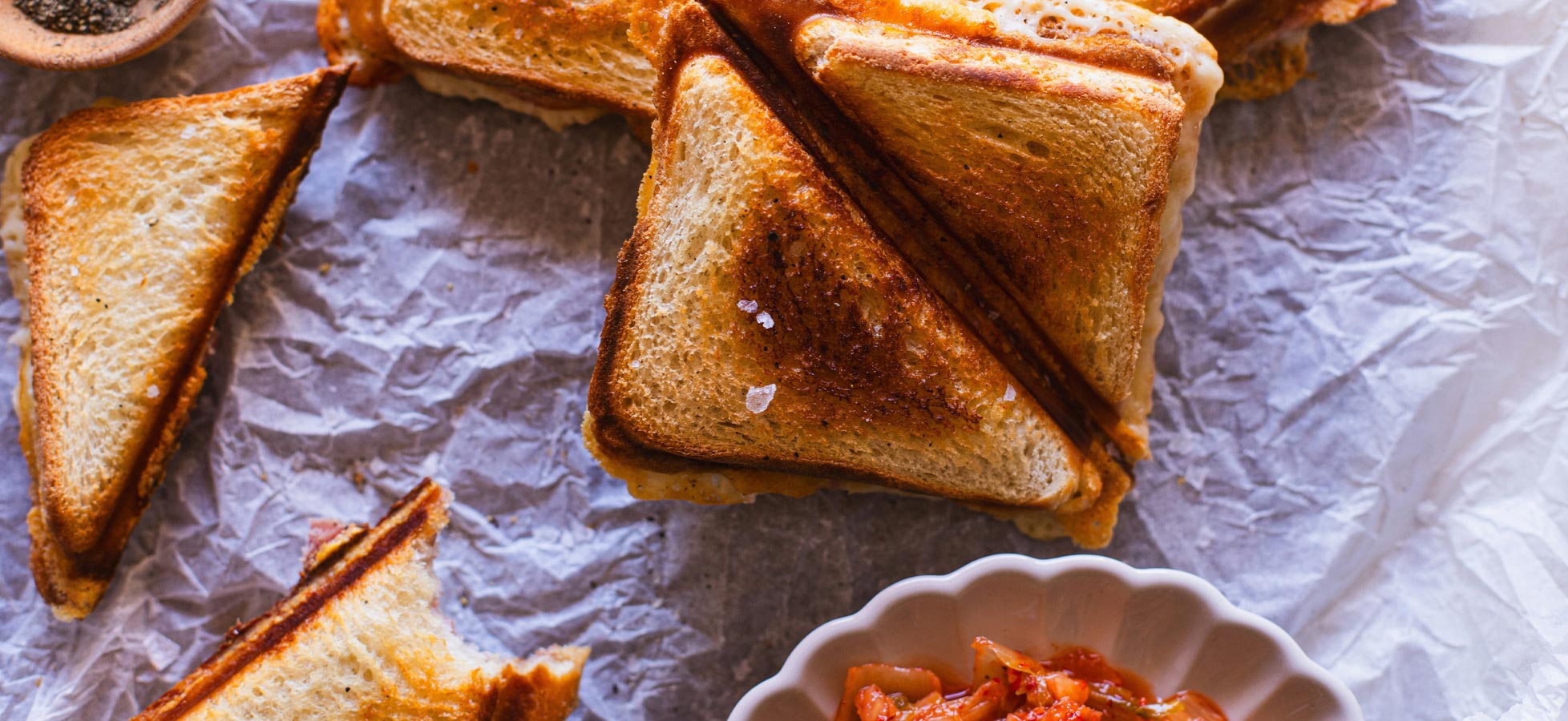 Auf einem Stück pergamentartigem Papier liegen mehrere goldbraune, dreieckige Sandwiches. Sie sind knusprig gebacken und leicht getoastet. Neben den Sandwiches befindet sich eine kleine, runde Schale mit eingelegtem Gemüse.