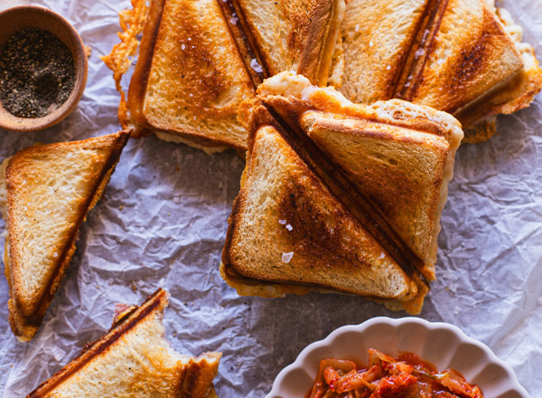 Eine Anordnung von goldbraun gerösteten Sandwiches mit geschmolzenem Käse, teilweise auf einem Stück Backpapier. Neben den Sandwiches befindet sich eine kleine Schüssel mit einer roten, würzigen Füllung und ein Holzschälchen mit schwarzem Pfeffer.