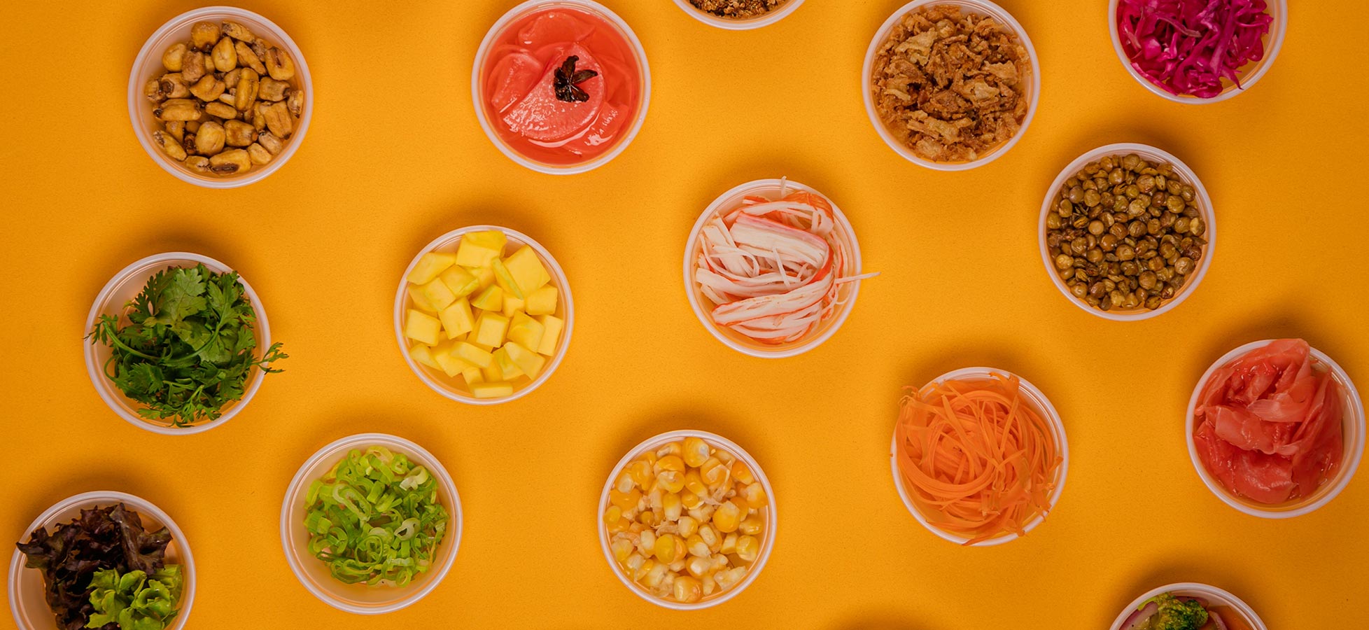 Eine Anordnung von kleinen Schalen mit verschiedenen Zutaten auf einem orangefarbenen Hintergrund. Die Schalen enthalten frische Kräuter, Obststücke, verschiedene Gemüse wie Karotten und darauf Zutaten wie Mais und Fisch. Die Vielfalt verdeutlicht eine bunte Auswahl an essbaren Zutaten.