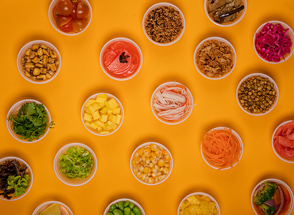 Das Bild zeigt eine Vielzahl von kleinen Schalen mit verschiedenen frischen Zutaten, darunter Gemüse, Obst und Snacks, auf einem leuchtend orangefarbenen Hintergrund. Die Schalen sind bunt angeordnet und enthalten unter anderem Tomaten, Mango, Karotten, Koriander und verschiedene Körner.