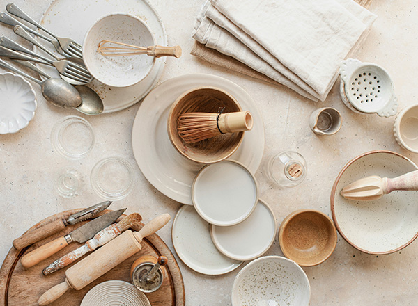 Eine Vielzahl von Küchenutensilien ist auf einem Tisch angeordnet. Dazu gehören Teller, Schalen, ein Schneebesen, Löffel und Ausstecher. Die Utensilien sind in unterschiedlichen Größen und Materialien gehalten. Ein helles, neutrales Farbschema dominiert die Szene.