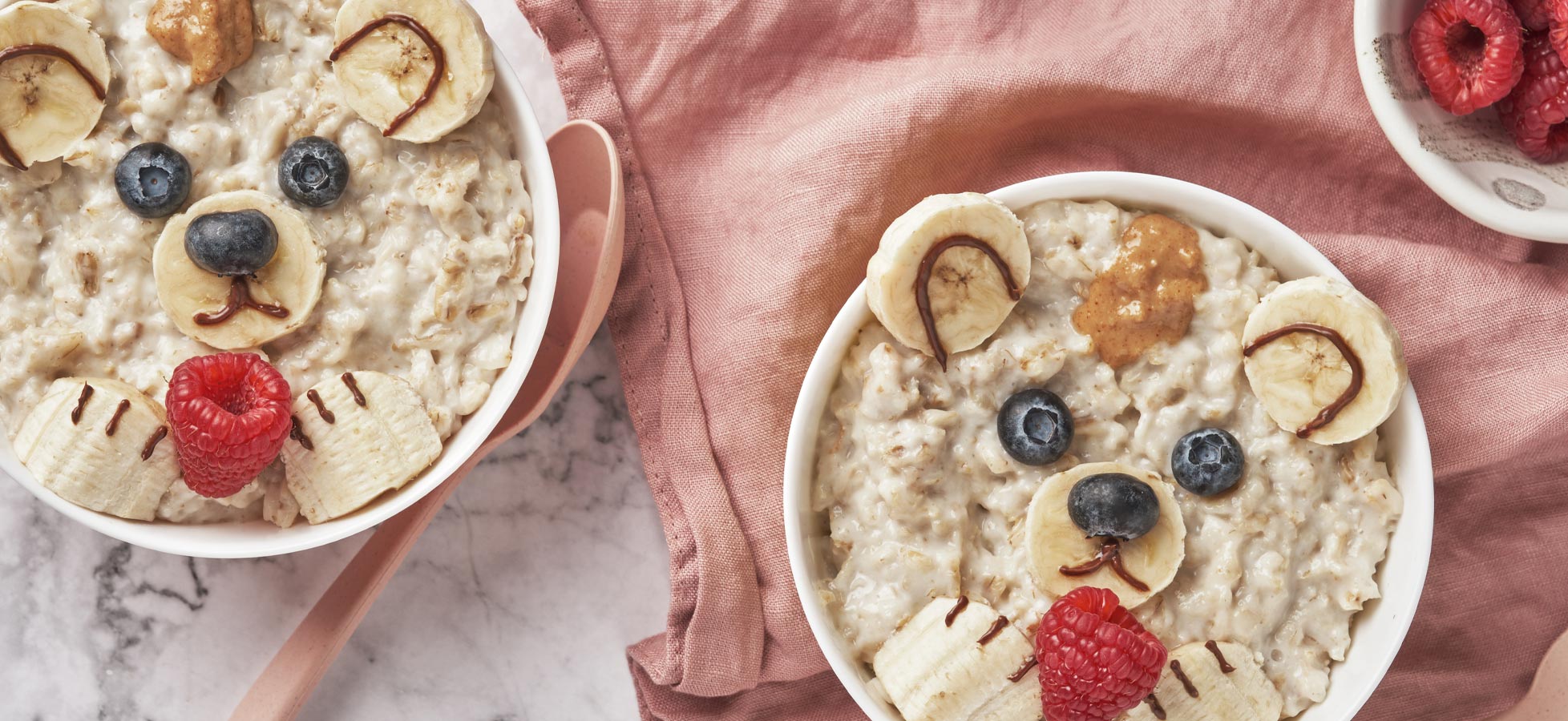 Zwei Schalen Haferbrei, kreativ dekoriert, um wie Bärenköpfe auszusehen. Die Gesichter bestehen aus Bananen, Heidelbeeren, Himbeeren und etwas Erdnussbutter. Die Schalen stehen auf einem marmorierten Tisch, umgeben von einem rosa Tuch und einem Löffel.
