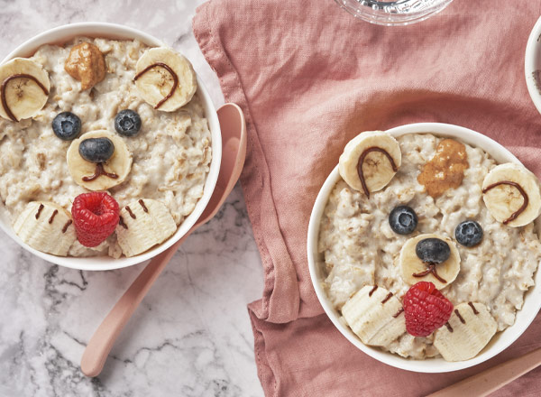 Zwei Schalen Haferbrei, kreativ dekoriert, um wie Bärenköpfe auszusehen. Die Gesichter bestehen aus Bananen, Heidelbeeren, Himbeeren und etwas Erdnussbutter. Die Schalen stehen auf einem marmorierten Tisch, umgeben von einem rosa Tuch und einem Löffel.