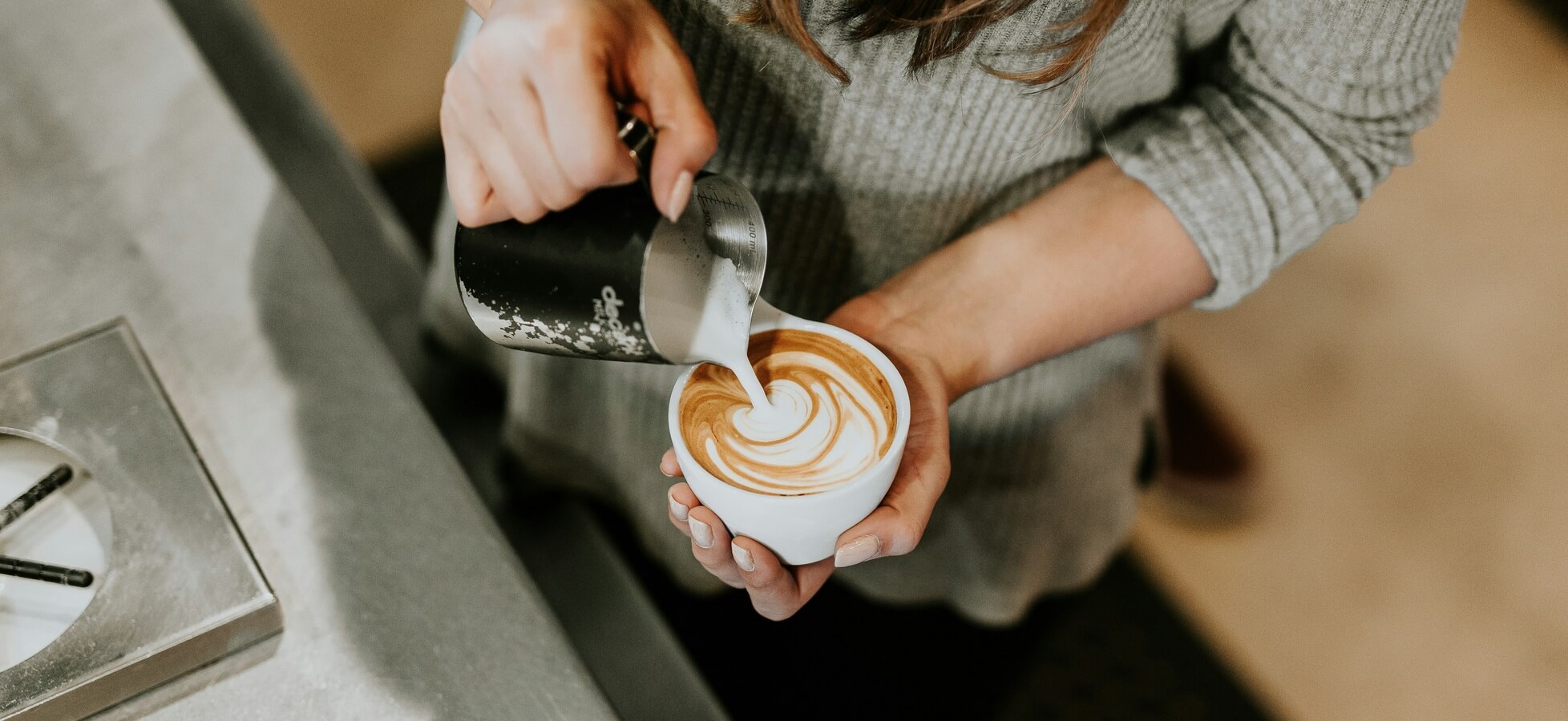 Eine Person gießt Milch aus einem Kännchen in eine Tasse und erstellt dabei kunstvolle Muster aus Schaum auf einem Kaffee. Die Tasse ist vor der Person positioniert, die in einem grauen Pullover gekleidet ist.