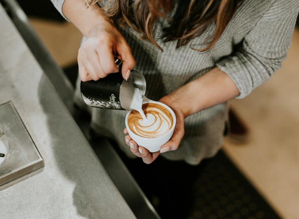 Eine Person in einem grauen Pullover gießt mit einem Kännchen Milch in eine Tasse Kaffee und kreiert dabei ein kunstvolles Muster aus Schaum. Hintergrund ist eine Café-Theke.