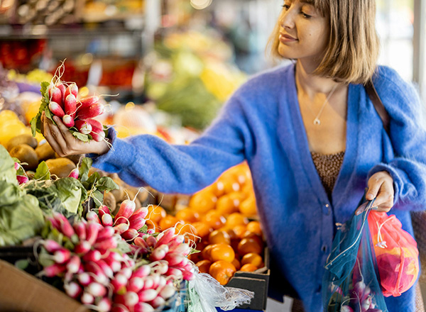 Eine junge Frau in einem blauen Pullover hält einen Bund Radieschen in der Hand, während sie auf einem Wochenmarkt steht. Um sie herum sind verschiedene Obst- und Gemüsesorten in Kisten angeordnet. Sie lächelt und scheint sich für die frischen Produkte zu interessieren.