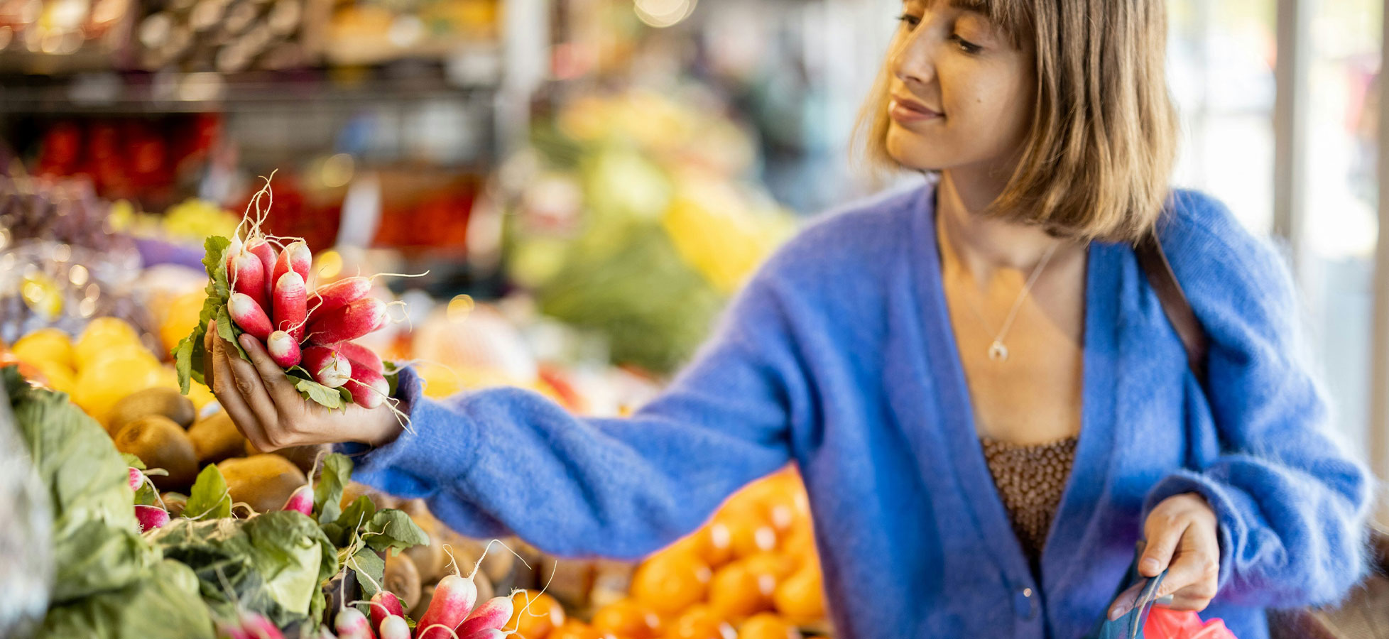 Eine Frau mit kurzen Haaren in einem blauen Pullover hält frische Radieschen in der Hand und betrachtet sie lächelnd. Im Hintergrund sind verschiedene Obst- und Gemüsesorten in einem Markt sichtbar.
